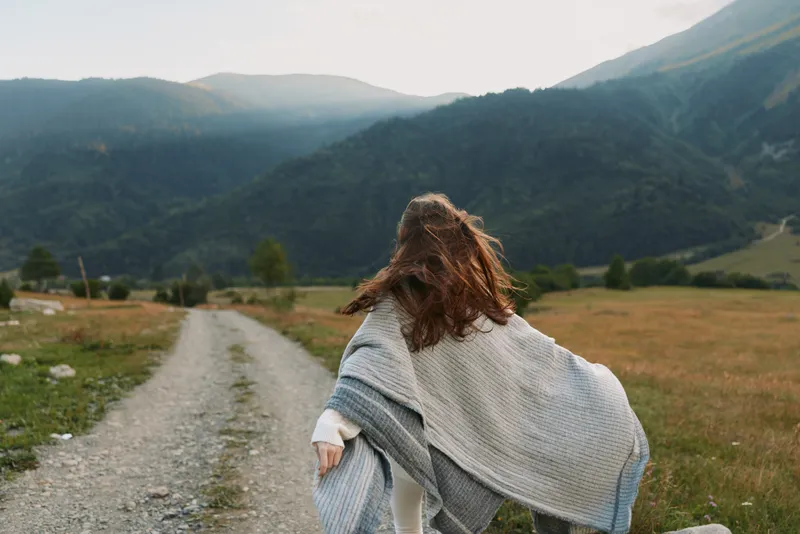A young woman in a light shawl walks along a rural gravel path toward distant mountains, embodying freedom and a calm outdoor lifestyle.