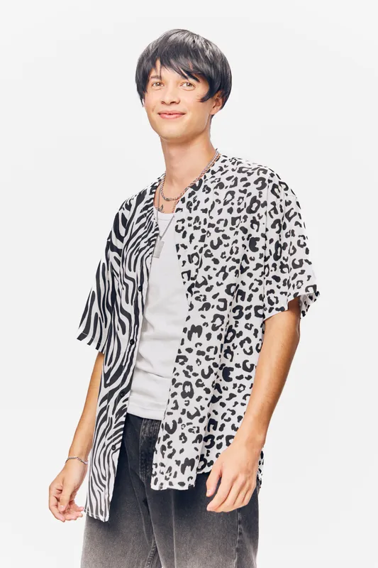 Young man wearing a stylish half leopard half zebra print shirt, casual white undershirt, and dark pants, standing against a clean white background with a relaxed smile.