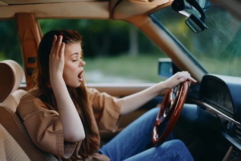 Frustrated young woman yawning while driving a vintage car in nature with brunette hair, casual clothing, and a relaxed expression...###