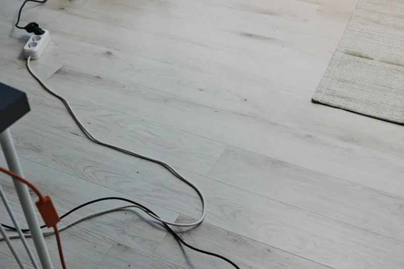 Tangled electrical cords on light wooden floor with a power strip near a rug edge, illustrating cable clutter and home office organization challenges.