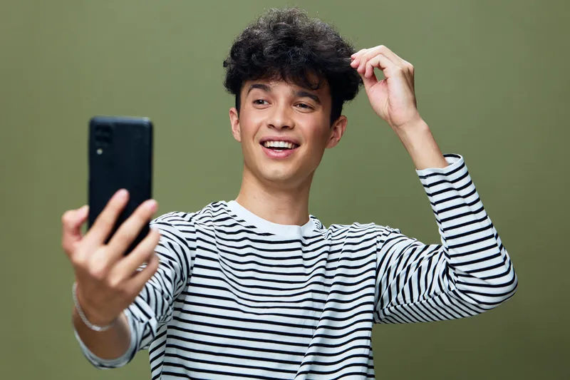 smiling young man taking selfie with smartphone, wearing striped shirt against green background, casual and joyful expression