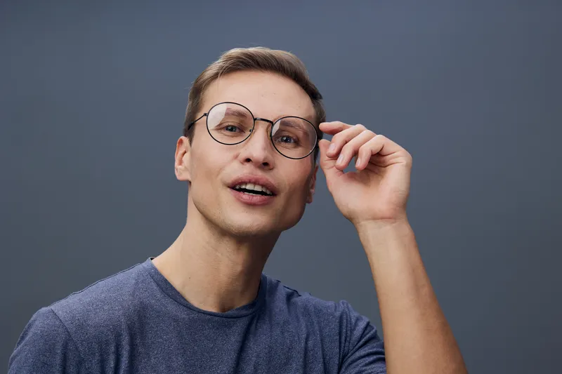 young caucasian man wearing glasses touch them with hand looking aside isolated on gray background casual style mental health and gadgets concept