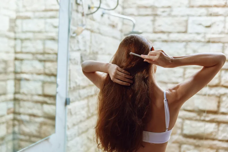 Woman with long brown hair brushing in bathroom near mirror, natural light, relaxed morning routine, white strap bra, calm atmosphere.