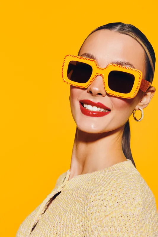 Smiling woman with bright red lipstick wears large orange sunglasses and yellow sweater against vibrant yellow background. Concept of fashion, summer style, and confident beauty.