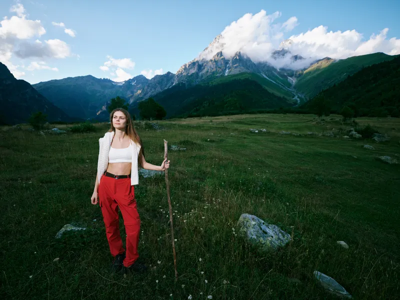 Woman standing in a mountain meadow with a walking stick, wearing red pants and a white top, expansive landscape with distant peaks and open outdoors