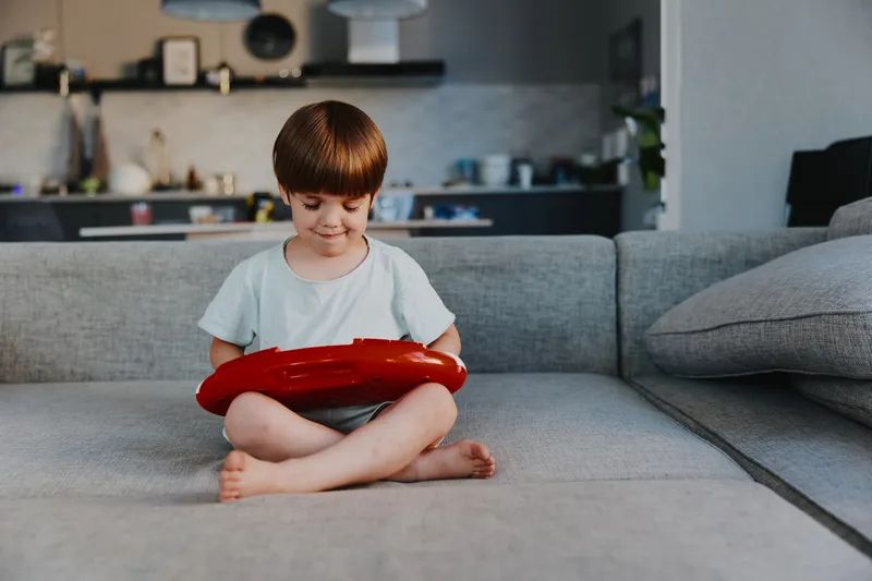 Joyful boy playing with a red toy instrument on a cozy sofa in a modern living room, enjoying his creative time and music exploration.