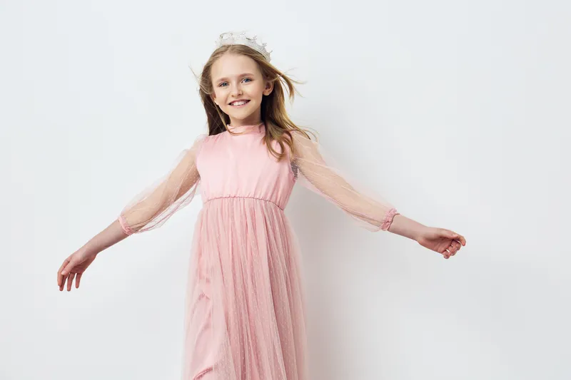 Happy girl in a pink dress with flowing sleeves, joyfully spinning with a smile, against a soft white background Perfect for children s fashion themes and joyful moments