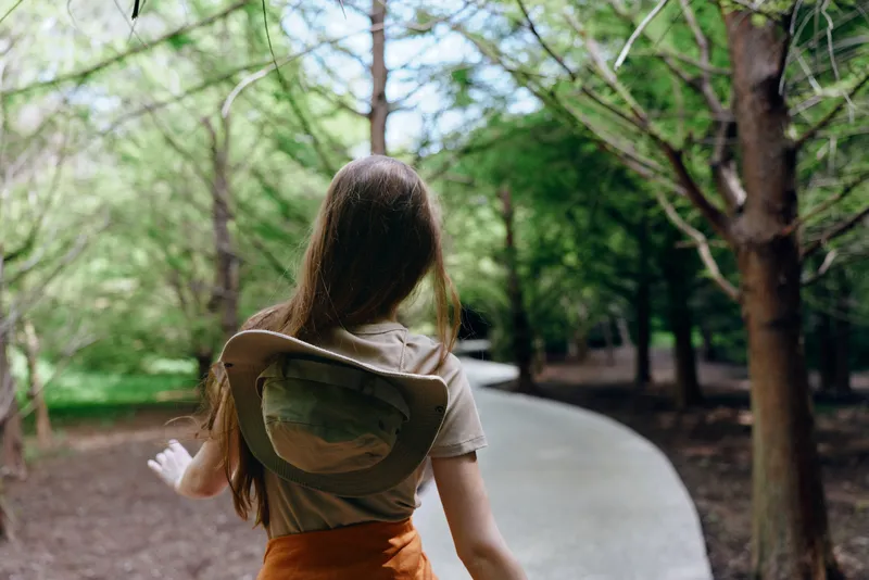 Woman walking on a shaded forest path with a backpack and hat, long hair flowing, enjoying nature among tall trees on a winding paved trail in a peaceful park setting.