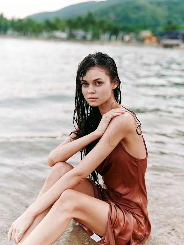 Young woman with wet hair sitting on the beach shore in a brown dress, looking thoughtfully with a serene natural background and soft daylight.