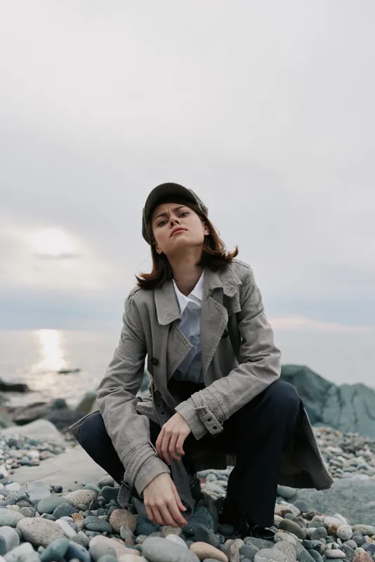 Young woman in a coat and cap squats on a rocky beach under cloudy sky near sea. Concept of fashion, casual style, confidence, and outdoor lifestyle with natural scenery.