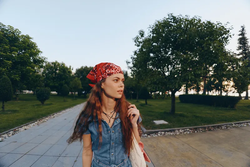Young woman with red hair wearing a bandana and denim dress walking outdoors in a green park with trees during daylight, looking thoughtful and casual.