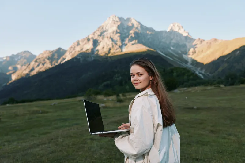 A cheerful young woman uses a laptop outdoors in a mountainous landscape, smiling softly as she works, blending technology with nature to create a calm, productive mood.
