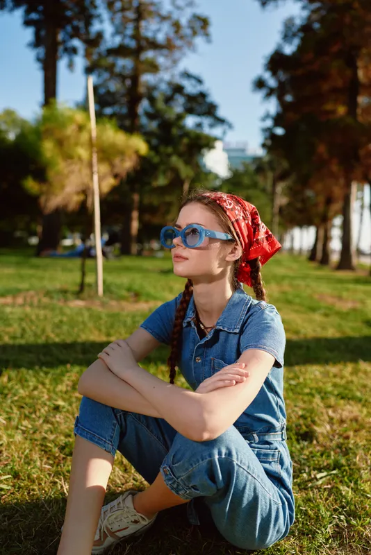 young woman wearing blue sunglasses and red bandana sitting on grass in sunny park with trees and cityscape in background