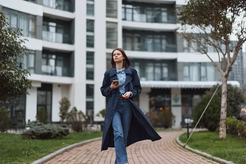 Young woman walks along a curved brick path in a city courtyard wearing a navy trench coat and blue denim, holding a smartphone, energetic stride, candid urban moment