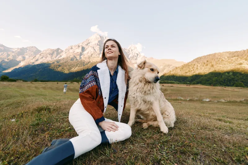Woman dog mountains meadow outdoors pet sitting nature landscape companion in a warm jacket, relaxed portrait of female and canine on grassy field enjoying alpine scenery, fresh air and freedom
