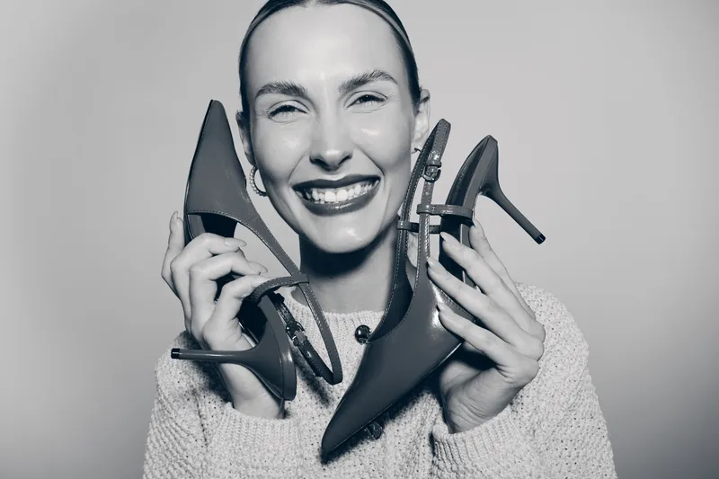A smiling Caucasian woman with light skin holds two pairs of high heel shoes near her face. The studio portrait shows fashion and footwear concept with a joyful expression.