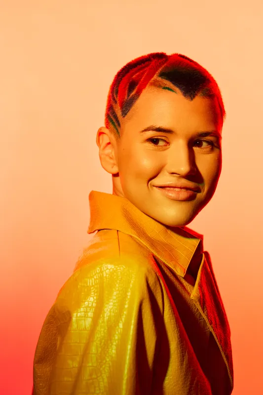 Woman with short braided hair wearing a yellow jacket, smiling subtly in warm orange lighting, showcasing modern style and youthful energy.