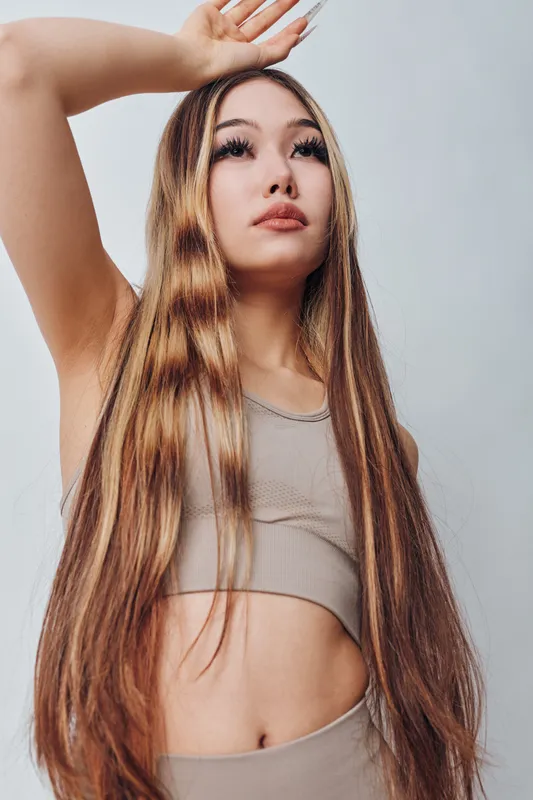 Young woman with long straight hair posing with raised arm in sleeveless crop top looking upwards against plain background in studio for fashion beauty portrait.