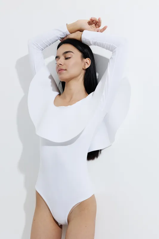 Woman sport fitness dressed in white bodysuit with unique ruffled design, posing with raised arms and closed eyes, showcasing modern style and calm expression in minimalistic studio.
