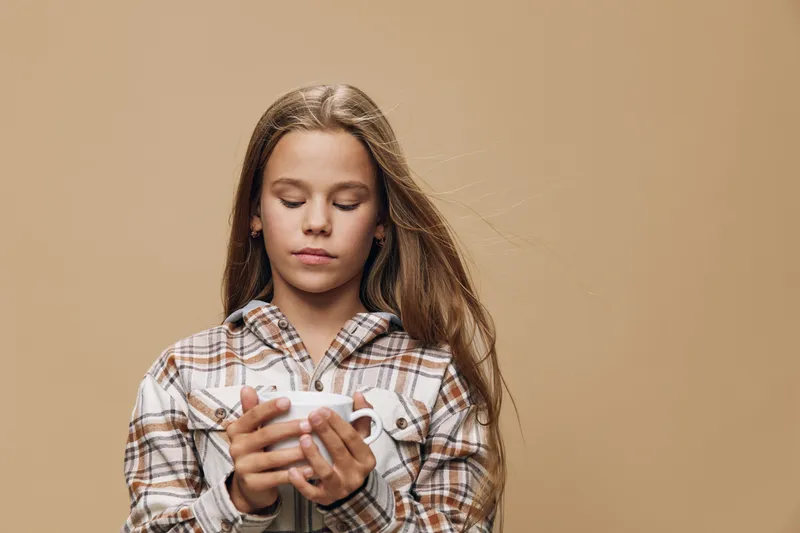 Girl with long hair wearing plaid shirt holding white cup with both hands standing against neutral background with calm and thoughtful expression.