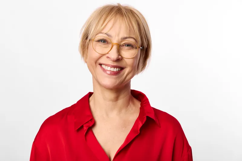 Woman smiling with glasses, senior blonde portrait in red studio shirt, friendly headshot, looking at camera, soft white background, positive mood, calm expression.