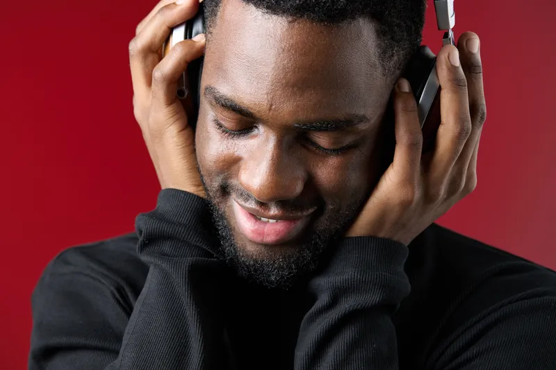 Young Black man enjoying music with headphones, smiling against a vibrant red background, showcasing emotions of joy and relaxation