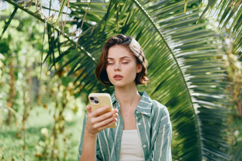 Young woman stands among lush tropical greenery, focused on a smartphone in her hand, wearing a casual striped shirt.