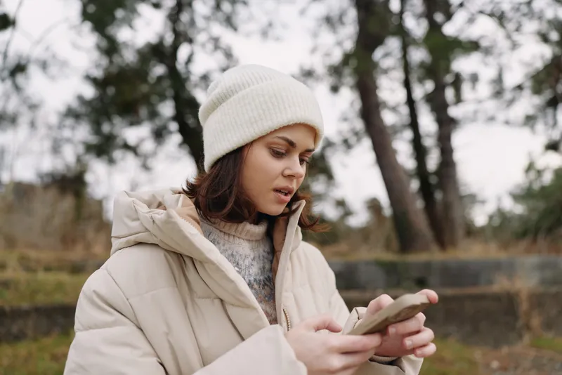 Winter woman using phone outdoors in a warm coat, calm park setting, focused on mobile communication and chilly weather for everyday life and work outdoors.