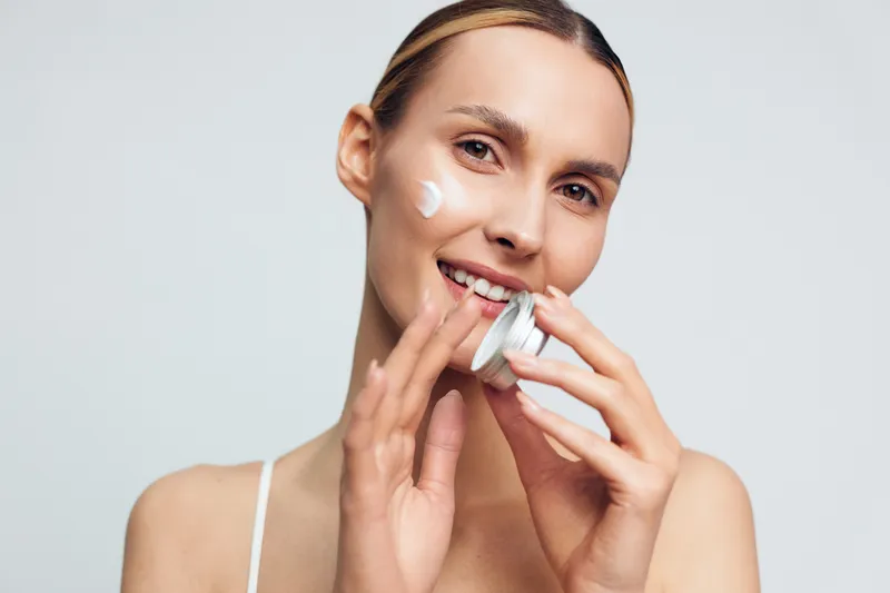 Young woman applying white face cream on cheek with fingers, smiling, with clear skin and tied hair, isolated on white background. Concept of skincare, beauty, and daily routine.