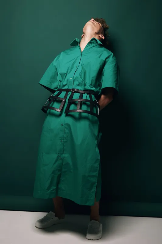 Man in green dress with short sleeves and black belt posing with head tilted back against dark green background in studio. Fashion, style and modern outfit with casual shoes and relaxed posture.