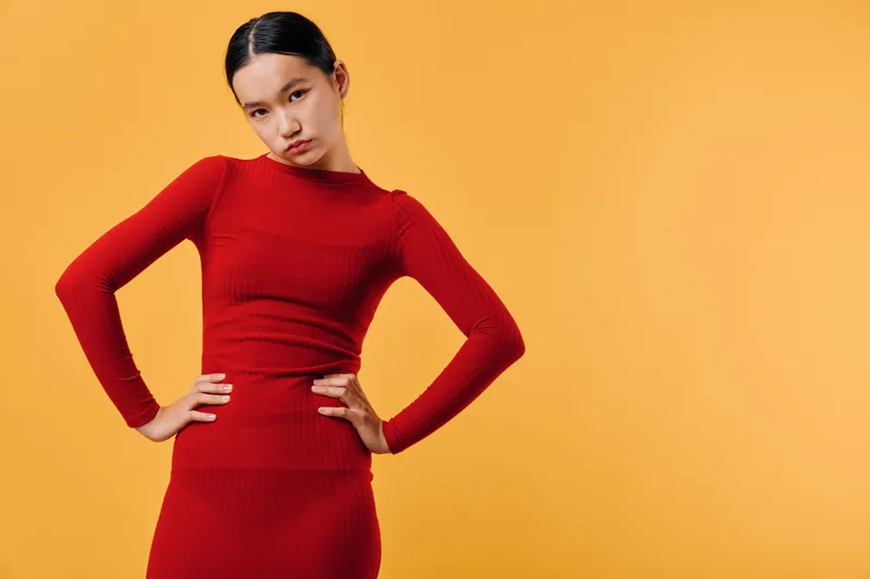 Woman posing in red dress with hands on hips against orange background. Fashion model with serious expression and sleek hairstyle in studio, showcasing stylish outfit and confident posture.
