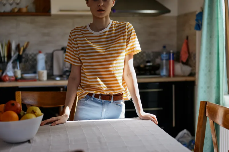 Casual woman stands in a home kitchen, leaning on a dining table, wearing an orange and white striped shirt and jeans. Natural light highlights everyday domestic life and a relaxed mood.