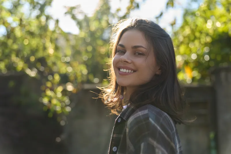 A smiling young woman poses outdoors in soft sunlight, wearing a casual plaid shirt. The warm natural backdrop suggests relaxed lifestyle, confidence, and everyday joy