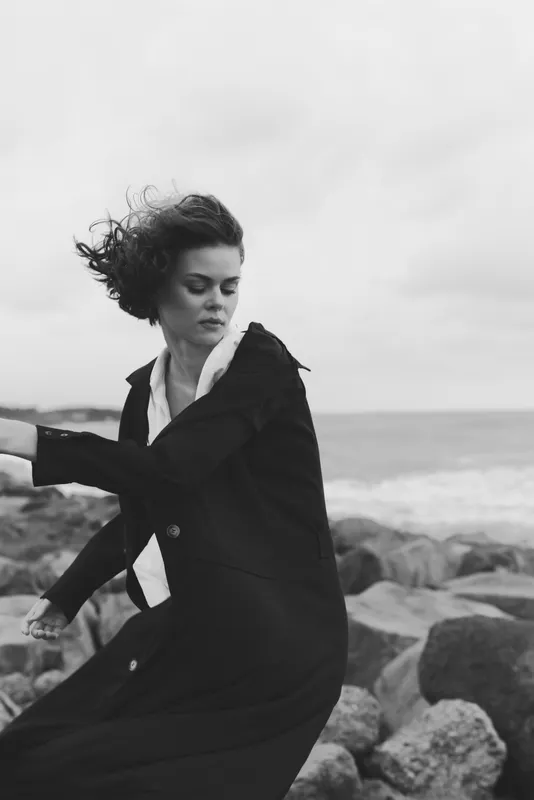 Windy seaside scene with a woman in a long coat near rough rocks, motion and focus captured as waves crash, dramatic monochrome fashion mood on a windy day by the shore.