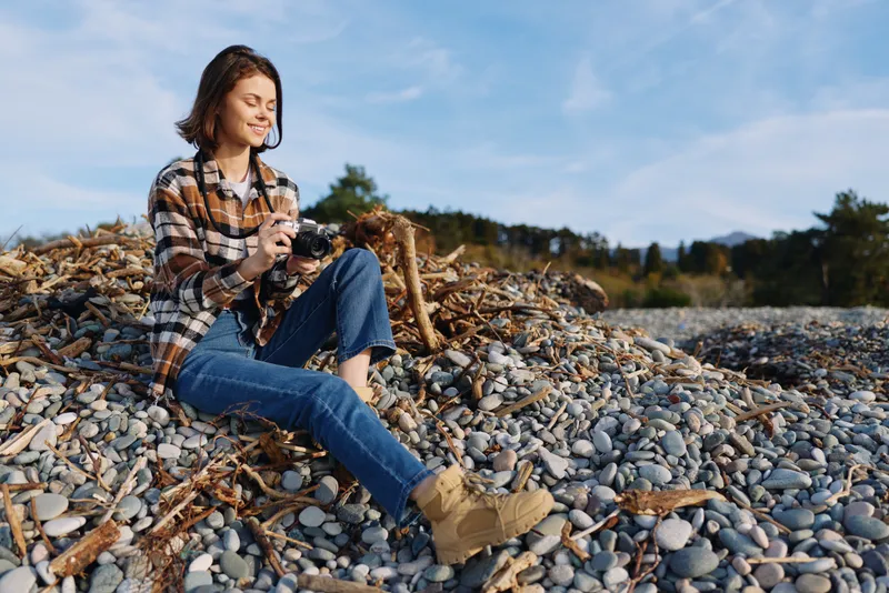 Woman sits on a pebbled shore outdoors, wearing a plaid shirt and jeans, using a camera or phone to capture a relaxed nature moment among driftwood and stones.