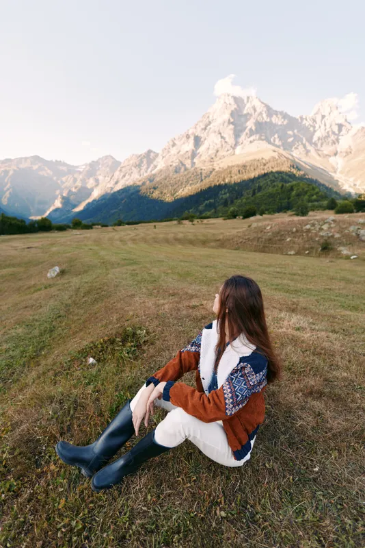 Woman sits on a meadow facing a mountain landscape during autumn travel, wearing cozy sweater and boots, enjoying serene scenery and wide grassy valley with soft sunlight.