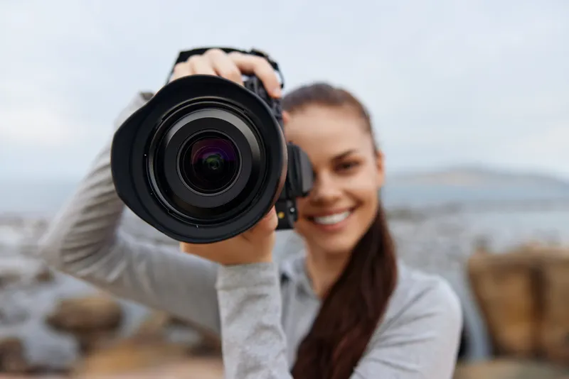 Smiling woman holding a camera at the beach in casual attire, expressing joy and enthusiasm for photography outdoors
