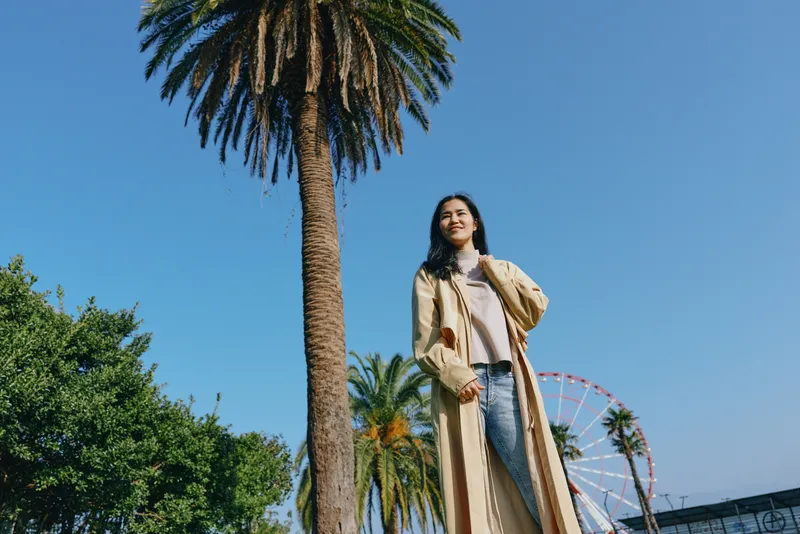 Woman in a beige coat stands beside palm trees under a clear blue sky, ferris wheel in the background, urban park scene, sunny day, chic style and fashion inspiration.