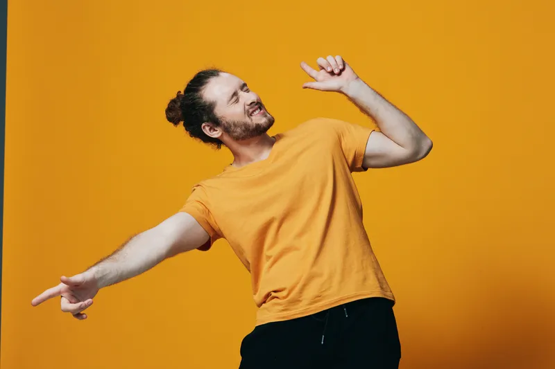 Cheerful young man in an orange t shirt dancing joyfully against a vibrant yellow background, showcasing expressions of happiness and carefree lifestyle