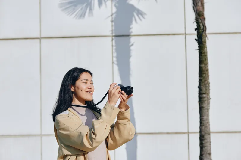 Photography of a smiling woman holding a camera outdoors beside a white wall with palm shadows, sunlight on her hair, casual sweater, capturing candid joy in a sunny urban or park setting