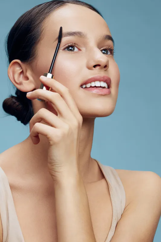 Young woman applying mascara with a joyful expression, showcasing beauty and self care against a soft blue background Perfect for cosmetic promotions