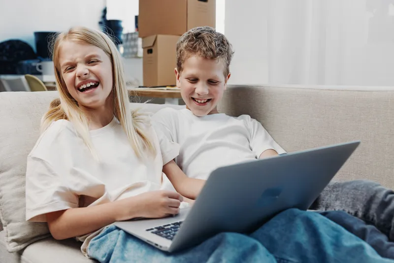 Two happy children sitting on a couch using a laptop, laughing and enjoying a relaxed moment at home. Concept of family, technology, and leisure time.
