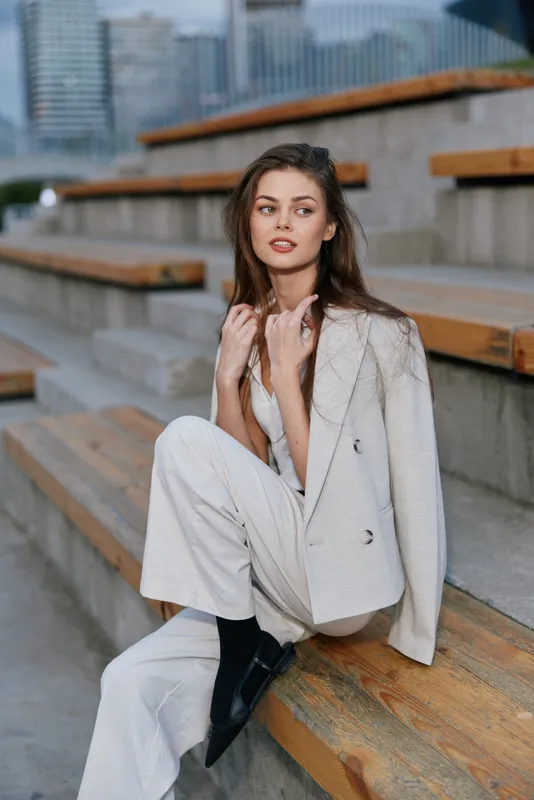 Elegant woman in light blazer and pants sitting on wooden steps outdoors in urban setting with thoughtful expression, fashion style, autumn season