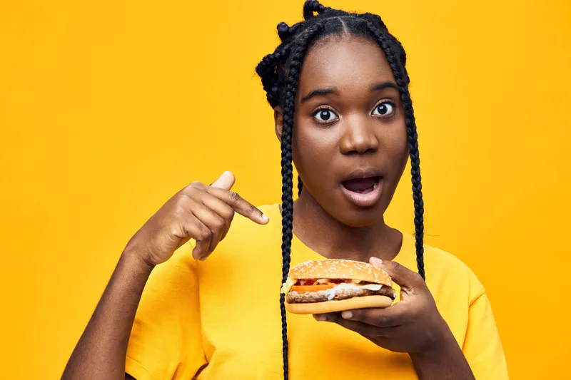 Excited young woman pointing at a delicious burger against a vibrant yellow background, showcasing fast food enthusiasm and joy