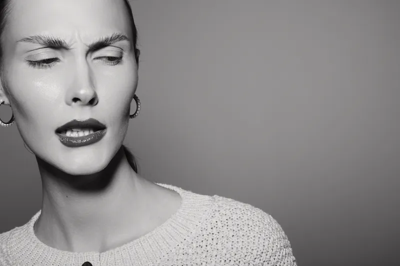 White woman with light skin wearing sweater looks confused or skeptical with furrowed brows and pursed lips in a studio setting with plain background.