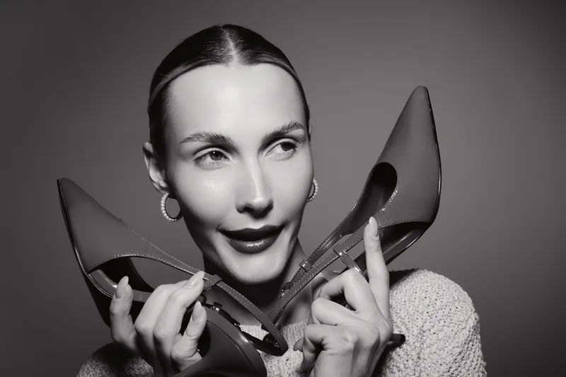 Young white woman with pale skin holds red high heels near face, smiling with playful expression in black and white studio portrait. Concept of fashion, style, and elegance.