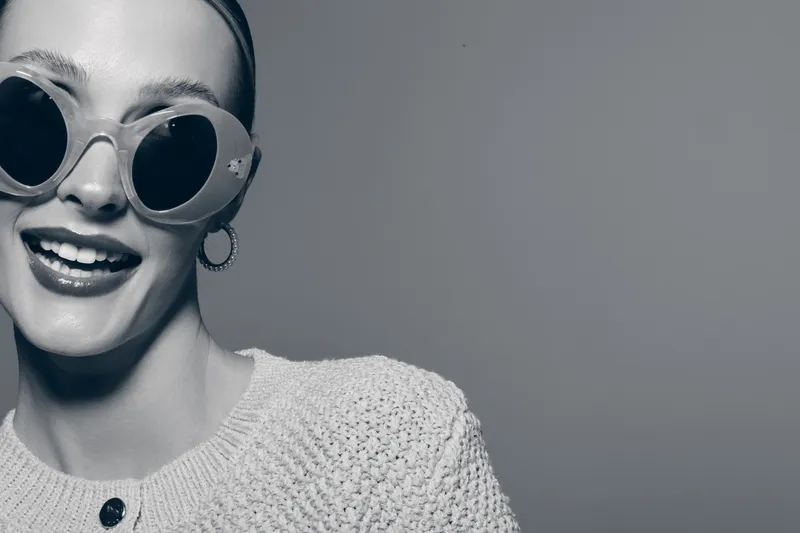 Smiling white woman with fair skin and sunglasses wearing knitted sweater poses cheerfully on gray background. Concept for fashion, lifestyle, happiness and casual style.