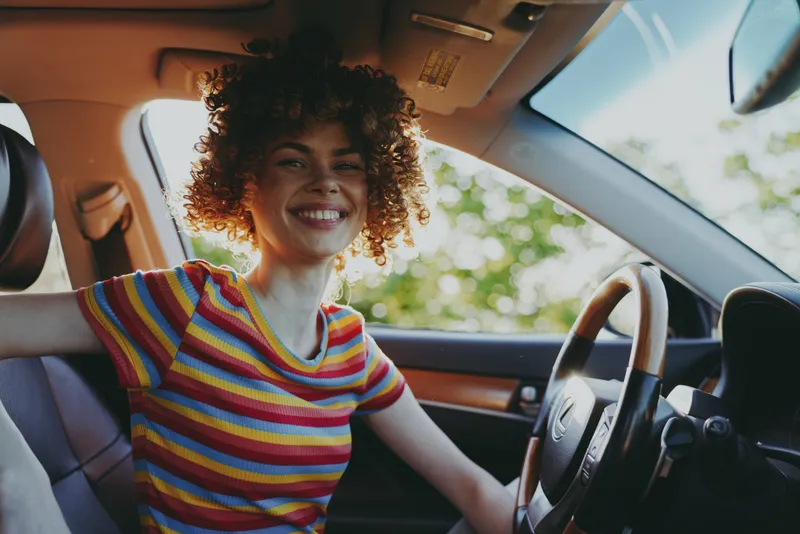 Cheerful woman inside a car, smiling with curly hair, striped rainbow shirt, sunlit interior, casual lifestyle moment, relaxed posture, natural colors, candid travel vibe