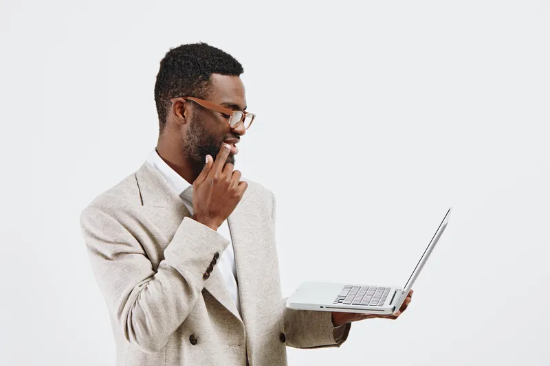 Thoughtful young man with glasses wearing beige blazer holding a laptop and touching his chin, standing against a plain white background in a studio setting. Business lifestyle concept