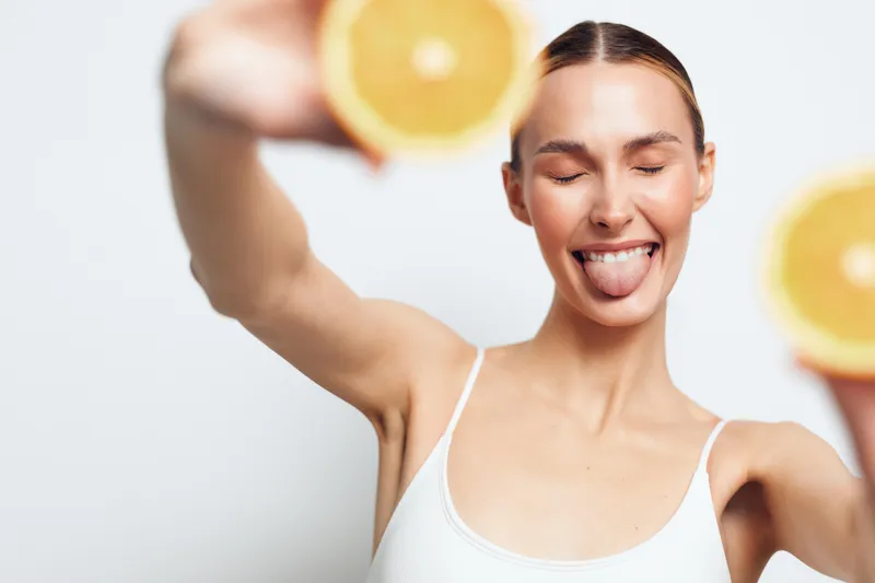 A happy young woman with closed eyes and tongue out holds two halves of a fresh orange in front of a white background. Concept for freshness, health, and summer vibes.